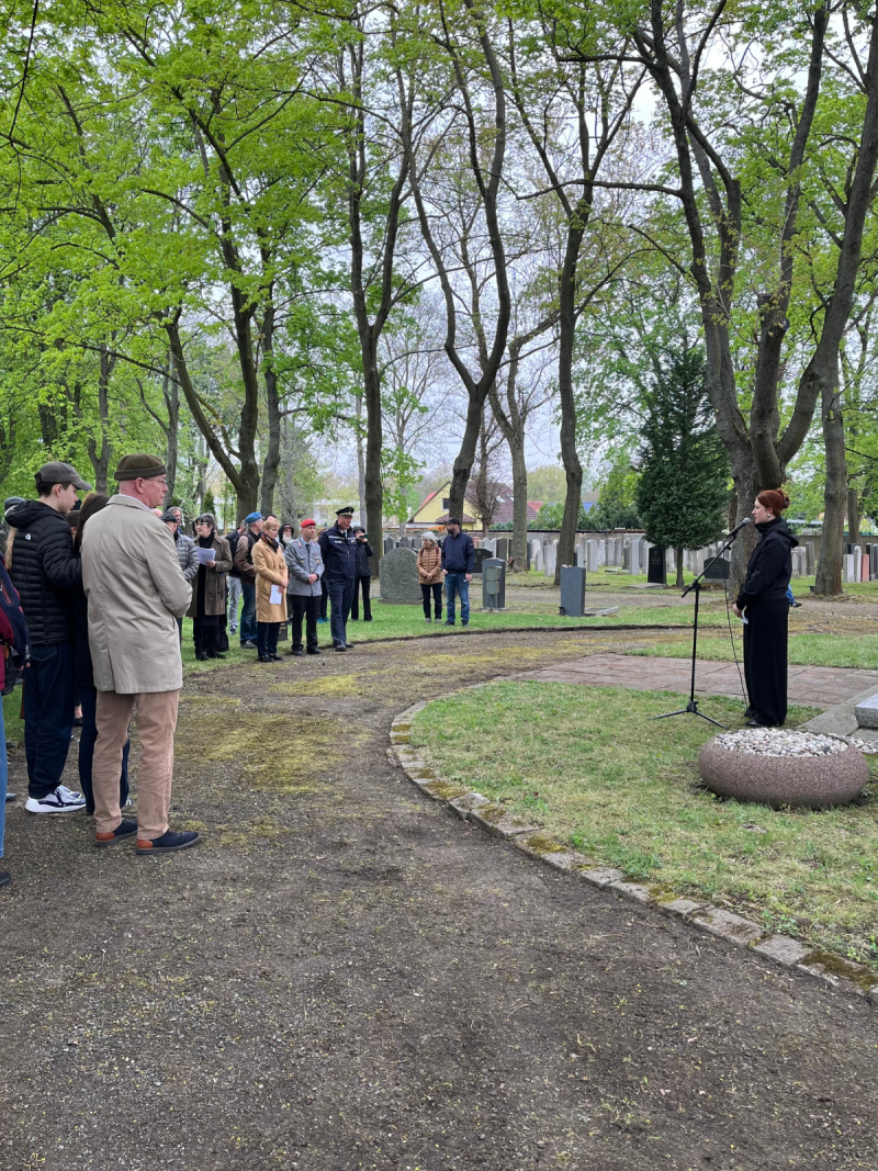 Eine Vetreterin des Jugendparlaments Leipzig spricht vor dem Holocaustdenkmal auf dem Neuen Israelitischen Friedhof Leipzig. Vor ihr steht eine Gruppe Zuhörender im Halbkreis
