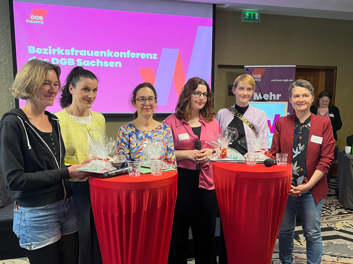 Claudia Maicher steht vor zwei Stehtischen. Links von ihr die gleichstellungspolitischen Sprecherinnen von CDU und Die Linke. Rechts von ihr Moderatorin Marsha Richarz, die gleichstellungspolitische Sprecherin der SPD und Daniela Kolbe vom DGB Sachsen. Auf den Stehtischen liegen Präsente für die Podiumsteilnehmerinnen.