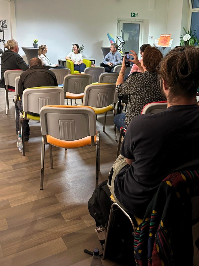 Claudia auf dem Podium zwichen zwei Personen, aus dem Publikum macht jemand Fotos