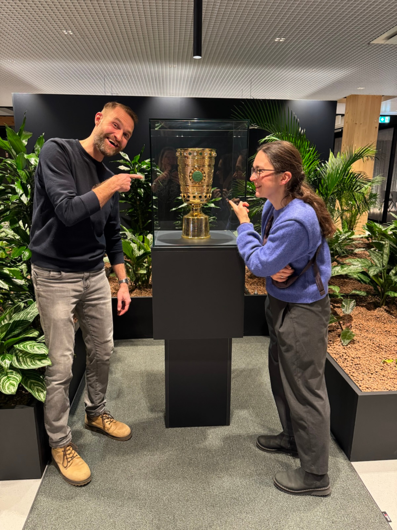 Claudia Maicher und Stadtrat Marvin Frommhold stehen neben einer Vitrine mit dem DFB-Pokal und zeigen auf ihn