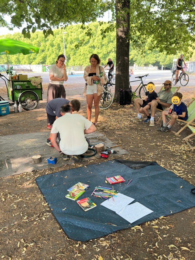 Ein Verantwortlicher der Geowerkstatt und ein Junge bohren zusammen Löcher in eine Scheibe aus einem Baum. Dabei werden sie von Umstehenden beobachtet.